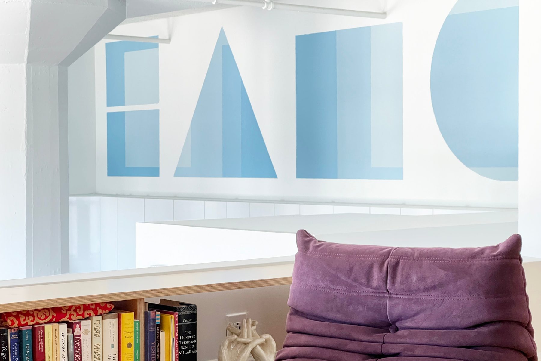 A tall white room with a large blue geometric wall painting and a low bookshelf